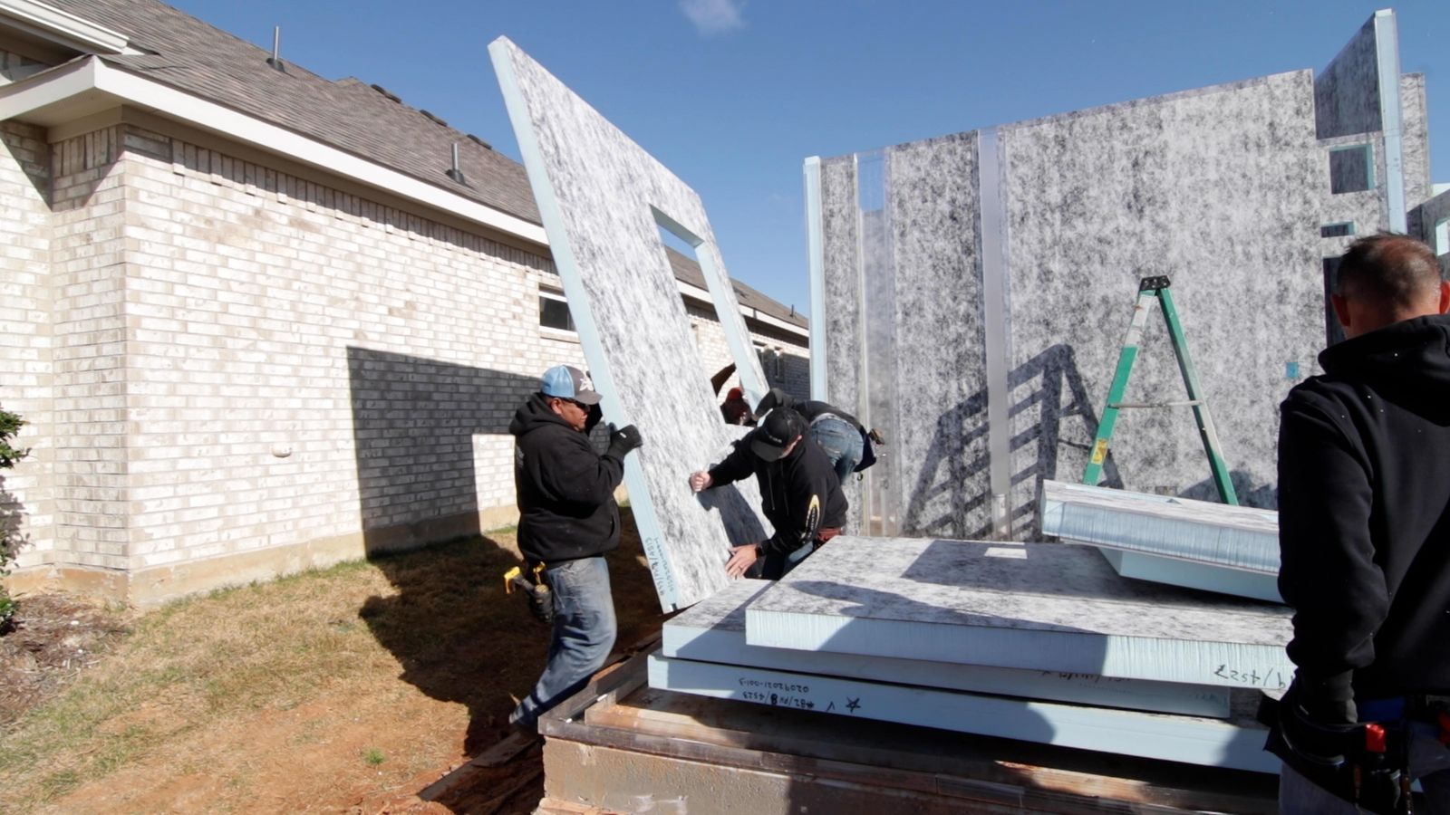 Crew guiding a tall wall panel into position beside a neighboring home